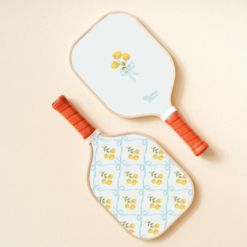 The-darling-effect's Marigold Lane Blue Pickleball Paddle features orange handles and lively designs: one with a yellow flower bouquet tied by a blue ribbon, the other with playful yellow flowers and blue ribbon patterns.