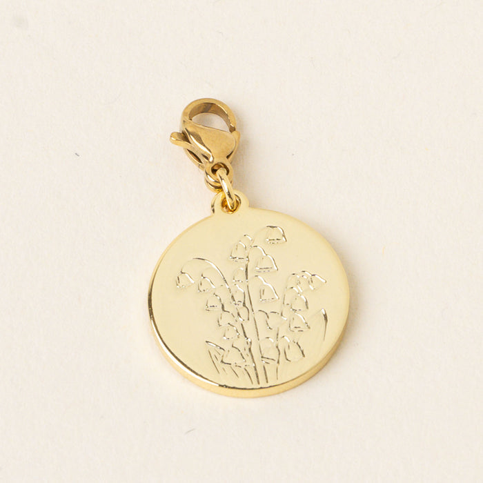 The May Jewelry Birth Flower Charm Gold by the-darling-effect features a gold round charm engraved with a lily of the valley, hanging from a gold clasp on a pale cream background—an ideal accessory for birth flower charm lovers.