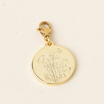 The May Jewelry Birth Flower Charm Gold by the-darling-effect features a gold round charm engraved with a lily of the valley, hanging from a gold clasp on a pale cream background—an ideal accessory for birth flower charm lovers.