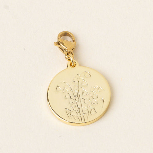 The May Jewelry Birth Flower Charm Gold by the-darling-effect features a gold round charm engraved with a lily of the valley, hanging from a gold clasp on a pale cream background—an ideal accessory for birth flower charm lovers.