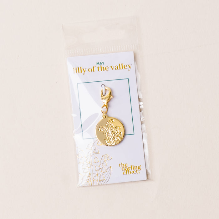 The-darling-effect's May Jewelry Birth Flower Charm Gold features an engraved lily of the valley and comes attached to a card with gold "darling effect" lettering, packaged in a clear plastic bag.