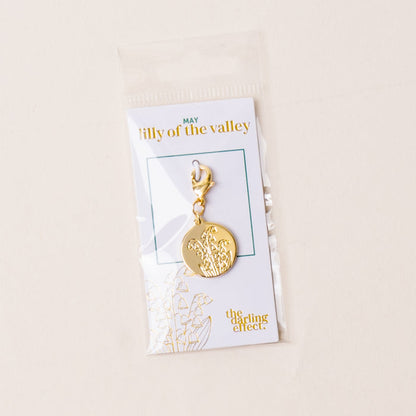 The-darling-effect's May Jewelry Birth Flower Charm Gold features an engraved lily of the valley and comes attached to a card with gold "darling effect" lettering, packaged in a clear plastic bag.