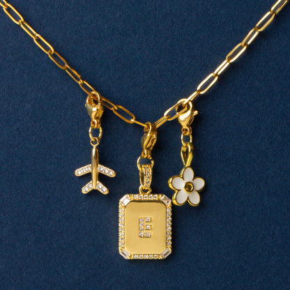 The Mia Paper Clip Chain Bracelet by the-darling-effect, displayed on a dark blue background, features a gold chain with three charms—a diamond-studded airplane, a diamond-encrusted initial E, and a white enamel flower—to elevate your personal style.