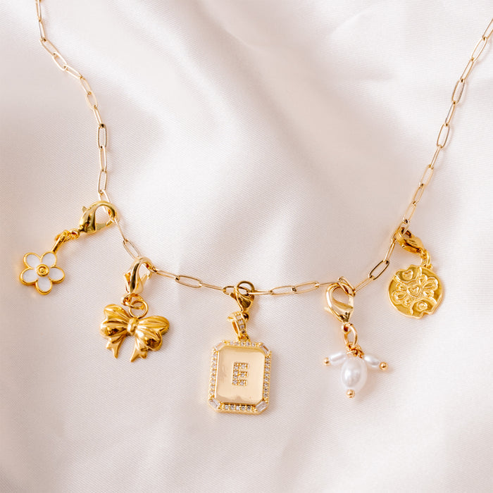 The Mia Paper Clip Chain Bracelet by the-darling-effect features five unique charms—a flower, a bow, a text pendant, a pearl cluster, and a round floral design—perfect for expressing your style and elegantly displayed on white fabric.
