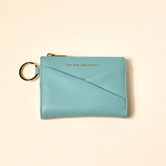 The Darling Effect’s Misty Blue Luxe Pocket Wallet is a small, light blue vegan leather wallet featuring a gold zipper, key ring, and "THE DARLING EFFECT" in gold text on the front, shown against a plain, light background.