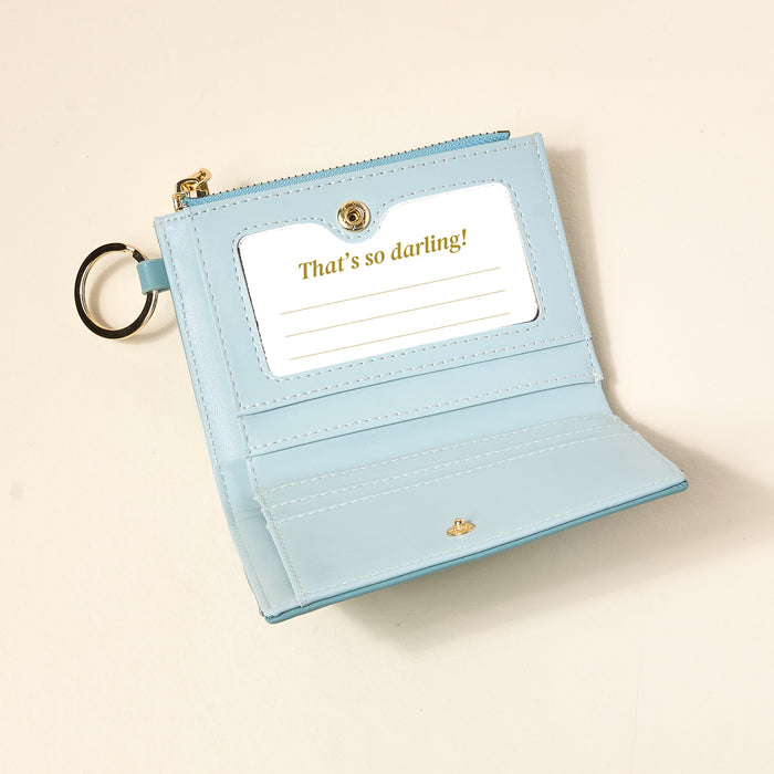 The Darling Effect's Misty Blue Luxe Pocket Wallet is a compact vegan leather wallet with a keyring, criss-cross pattern, and card slot displaying a gold “Thats so darling!” card, shown on a light beige surface.