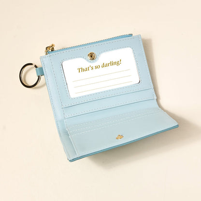 The Darling Effect's Misty Blue Luxe Pocket Wallet is a compact vegan leather wallet with a keyring, criss-cross pattern, and card slot displaying a gold “Thats so darling!” card, shown on a light beige surface.