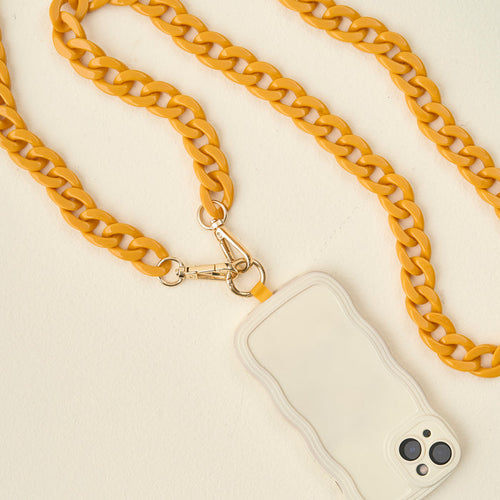 A smartphone with a beige wavy-edged case is attached to a thick, yellow crossbody chain strap with gold clasps, creating a fashionable accessory on a cream-colored surface.