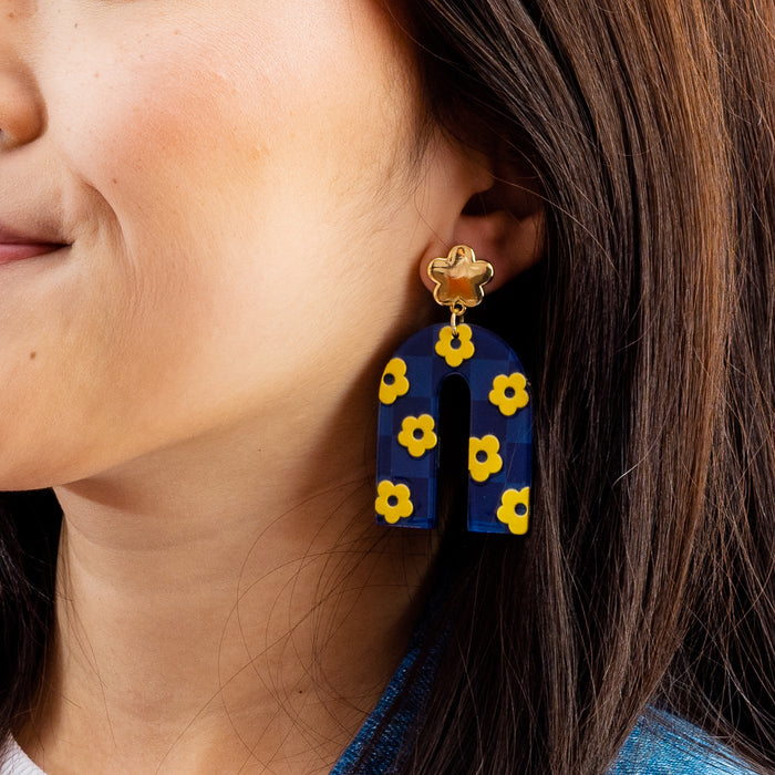 A close-up of a woman in The Darling Effect’s Navy Yellow Game Day Acrylic Earrings—large, arch-shaped navy acrylic with yellow flower details and a gold-flower stud—her brown hair down, smiling. The perfect accessory to show your school colors!.