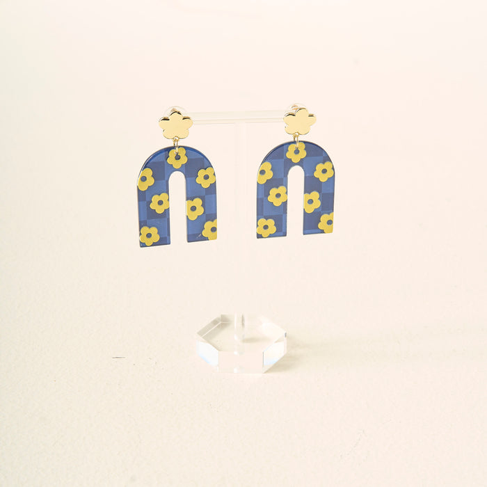 Show off your school colors with The Darling Effect’s Navy Yellow Game Day Acrylic Earrings, featuring gold-colored flower studs and blue arch dangles with yellow flowers, displayed on a clear stand against a light background.