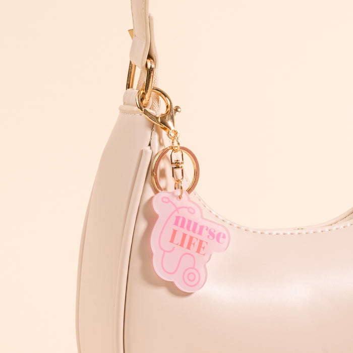 A close-up of a white handbag with the-darling-effect's Nurse Life Acrylic Keychain attached, featuring a pink and white medical badge charm and bold "nurse LIFE" lettering.