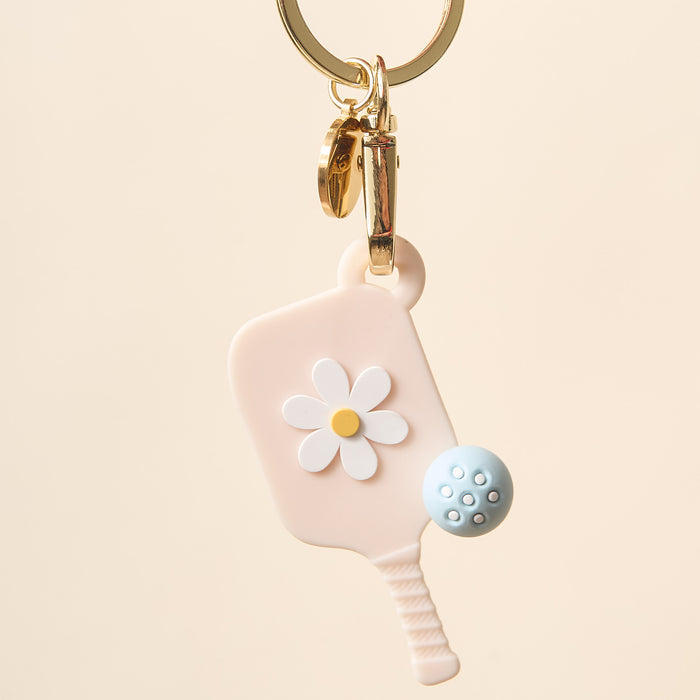 The Darling Effect's Oat Pickleball Paddle Keychain features a pastel pink mini paddle with a white daisy and blue ball on a gold keyring—an adorable pickleball gift.