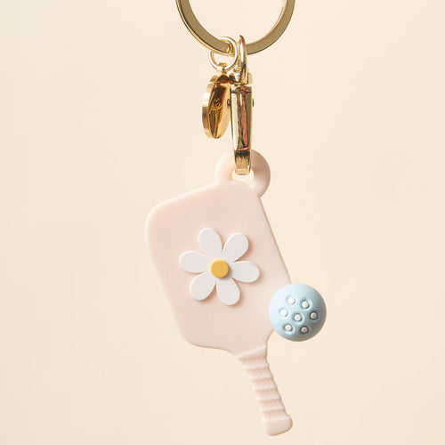 The Darling Effect's Oat Pickleball Paddle Keychain features a pastel pink mini paddle with a white daisy and blue ball on a gold keyring—an adorable pickleball gift.