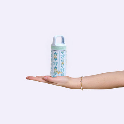 A hand with a bracelet holds The Darling Effect's Oh So Fancy Blue 6 oz Lil' Bitty Bottle—white and light blue with yellow and blue floral patterns—making it the perfect travel companion against a plain light background.