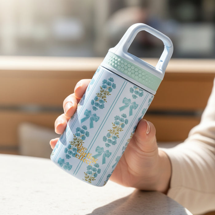 A person holds The Darling Effect's Oh So Fancy Blue 6 oz Lil' Bitty Bottle, featuring floral and bow patterns with a mint green lid, over a white table—perfect for on-the-go hydration.