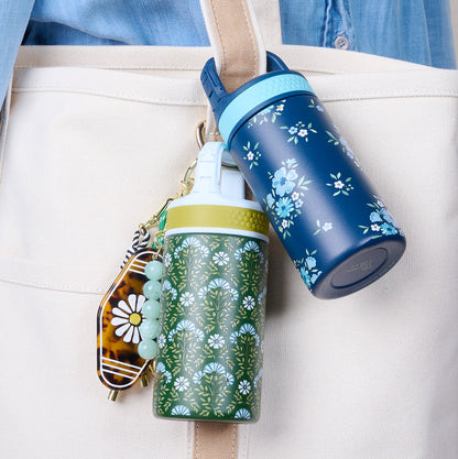 Two Book Shelfie 6 oz Lil' Bitty Bottles by The Darling Effect—a navy blue with blue flowers and a green with white daisies—clip onto a cream tote with a keychain charm, making stylish, floral travel companions.