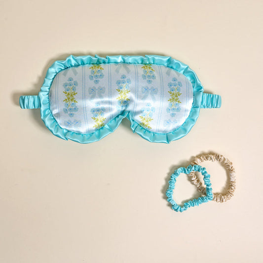 The Darling Effect’s Oh So Fancy Blue Beauty Sleep Mask, featuring yellow florals and ruffled edges, is displayed beside blue and beige satin scrunchies on a light beige surface—an elegant addition to your nightly routine.