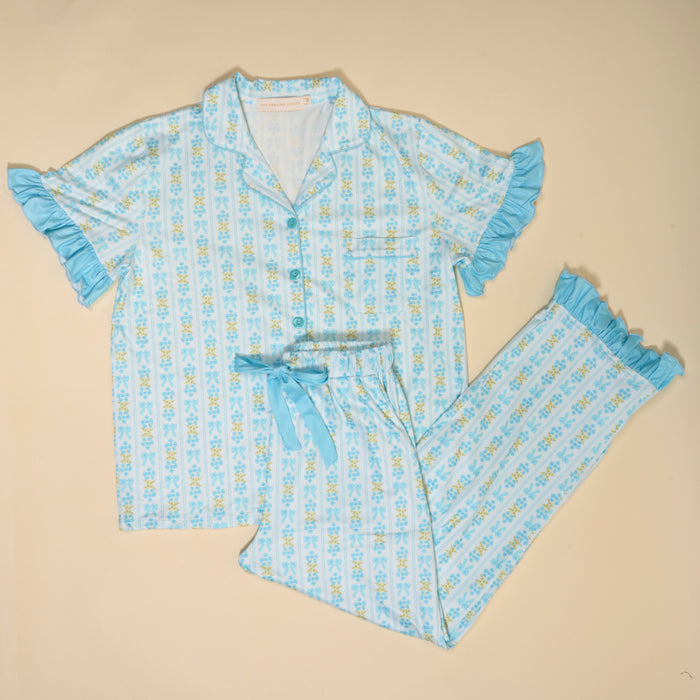 The Darling Effect Oh So Fancy Blue Slumber Party Pajama Pants Set features ultra-soft, light blue fabric with yellow and blue florals, ruffled edges, a short-sleeve button-up top, and long pants laid flat on a beige background.