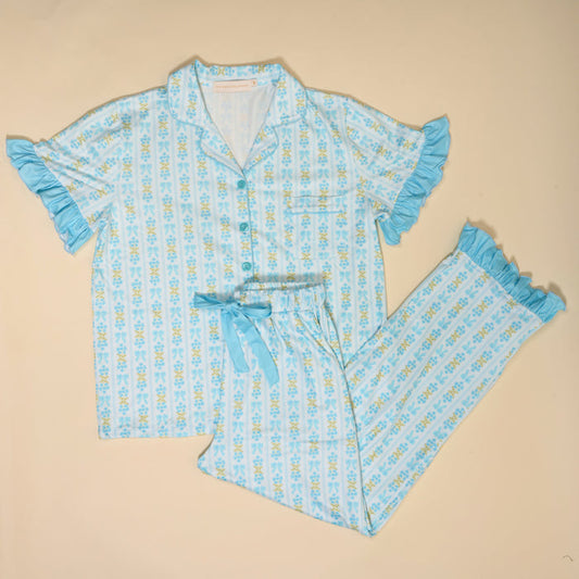 The Darling Effect Oh So Fancy Blue Slumber Party Pajama Pants Set features ultra-soft, light blue fabric with yellow and blue florals, ruffled edges, a short-sleeve button-up top, and long pants laid flat on a beige background.
