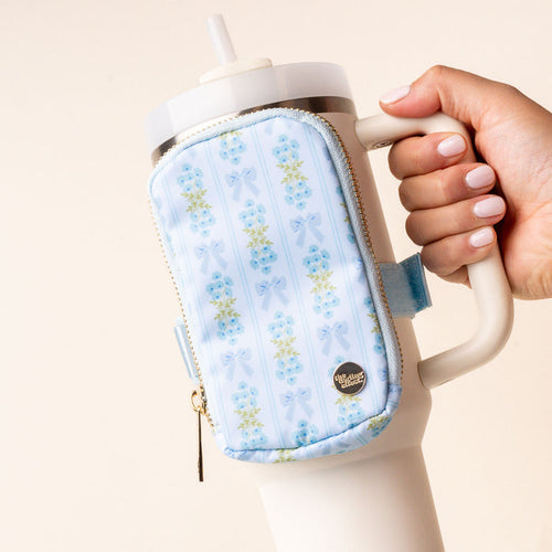 The Oh So Fancy Blue Tumbler Fanny Pack by the-darling-effect, featuring a floral and bow design, is attached to a white travel mug held by a hand with light pink nail polish.