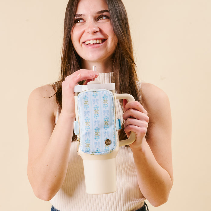 A smiling woman holds a cream tumbler with a metal straw and the Oh So Fancy Blue Tumbler Fanny Pack by the-darling-effect, featuring blue florals and water-resistant material, set against a beige background.