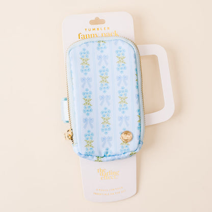 The Oh So Fancy Blue Tumbler Fanny Pack by the-darling-effect features a stylish light blue floral and bow pattern, gold zipper and charm, is water resistant, and comes on a white card with the-darling-effect logo.