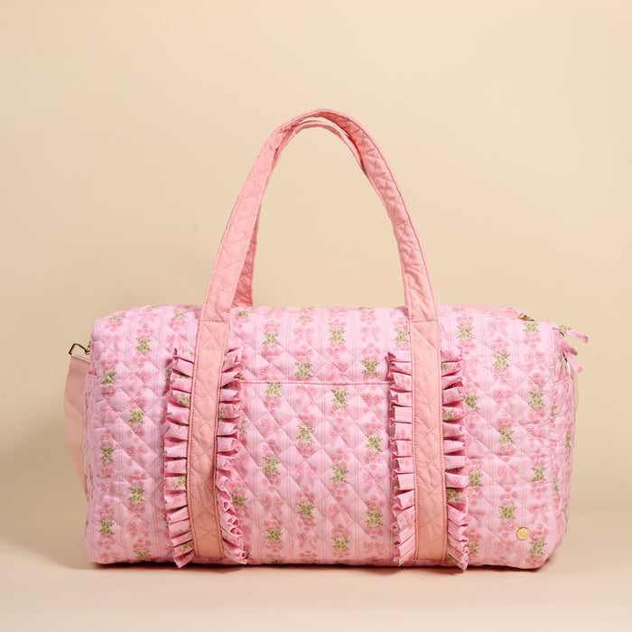 The Darling Effect's Oh So Fancy Pink On-the-Go Quilted Weekender Bag features floral patterns, ruffled trim, and two handles—a stylish, spacious choice for your next getaway. Set against a beige background.