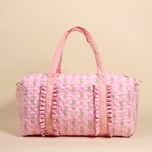 The Darling Effect's Oh So Fancy Pink On-the-Go Quilted Weekender Bag features floral patterns, ruffled trim, and two handles—a stylish, spacious choice for your next getaway. Set against a beige background.