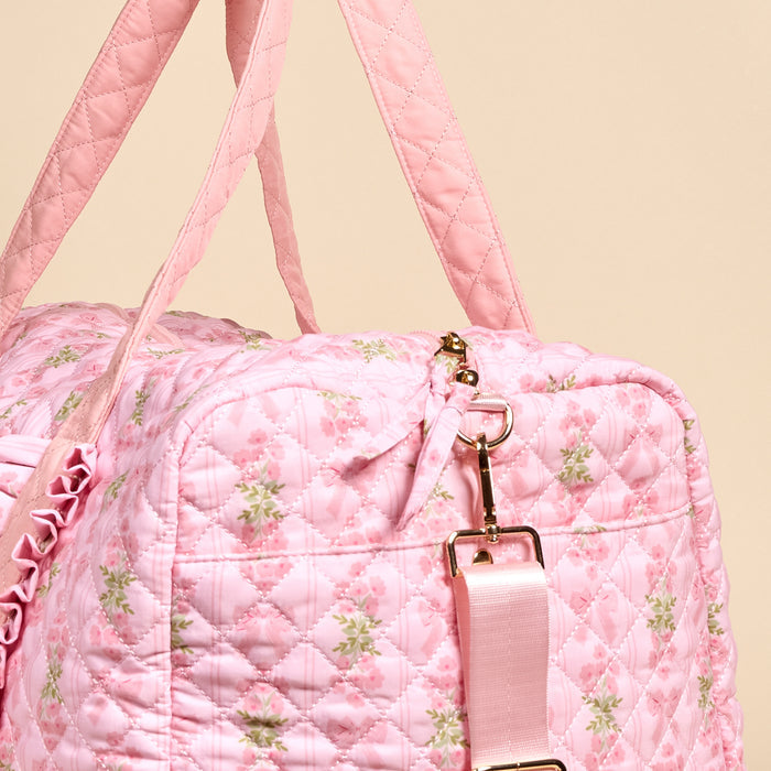 A close-up of The Darling Effect's Oh So Fancy Pink On-the-Go Quilted Weekender Bag features a pink floral pattern, gold hardware, pink handles, and an adjustable shoulder strap against a neutral background.