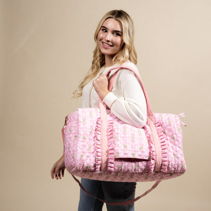 A smiling woman with long blonde hair carries The Darling Effect's Oh So Fancy Pink On-the-Go Quilted Weekender Bag on her shoulder while wearing a white sweater and jeans, standing against a beige background.
