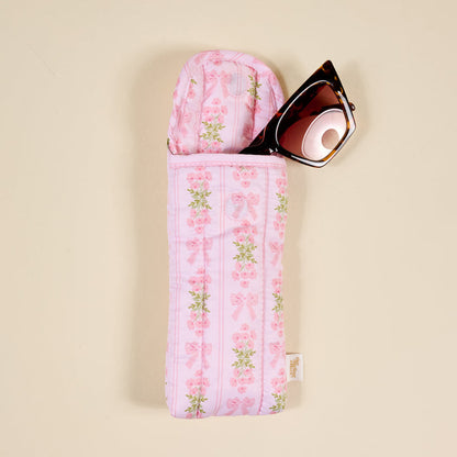 A pair of sunglasses partially tucked into The Darling Effect's Oh So Fancy Pink Puffy Sunglasses Case, featuring a floral and bow pattern with a magnetic flap closure, resting on a beige surface.