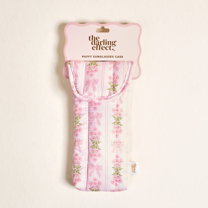 The Darling Effect's Oh So Fancy Pink Puffy Sunglasses Case features a pink and white quilted design with bow and flower accents, a magnetic flap closure, side tag, and comes in packaging labeled "PUFFY SUNGLASSES CASE.