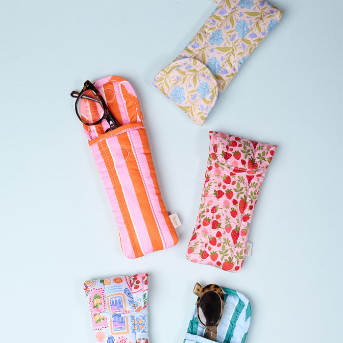 Five Oh So Fancy Pink Puffy Sunglasses Cases by The Darling Effect—some quilted and vibrantly patterned with magnetic flap closures—are arranged on a light blue surface. Two machine-washable cases have sunglasses partially inside.