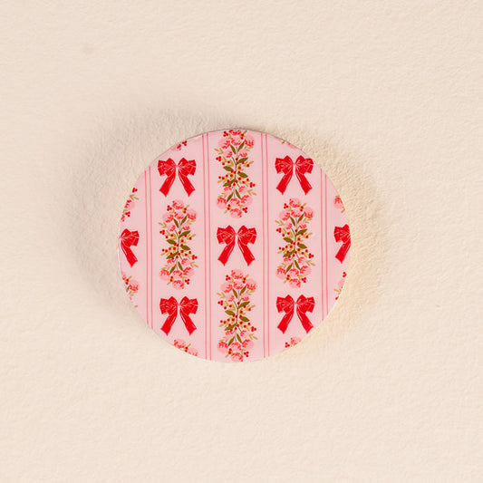 The Oh So Merry Wood Enamel Coaster by The Darling Effect features a pink background with vertical stripes of red bows and small pink flowers, and sits on a textured off-white surface.