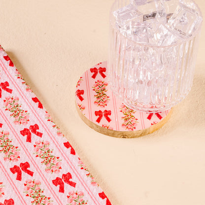 A clear glass with ice rests on The Darling Effect's Oh So Merry Wood Enamel Coaster, adorned with red bows and flowers, beside fabric featuring a matching red bow and flower design on a light surface.