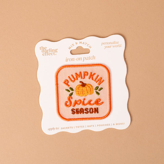 The Darling Effect’s On Patch-Pumpkin Spice Season Mix & Match Iron is a square iron-on patch featuring an orange pumpkin and the phrase “Pumpkin Spice Season” embroidered on it, displayed on beige packaging. Perfect for customizing clothing or accessories, this patch adds a cozy touch to any colorful collection.