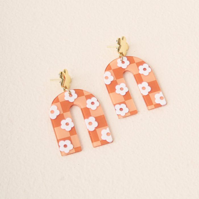 The Darling Effect's Orange Game Day Acrylic Earrings feature arch-shaped acrylic with an orange and white checkered pattern, white daisies, and gold rabbit posts—perfect for game day. Displayed on a light beige surface.