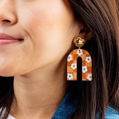 A woman with straight brown hair wears The Darling Effect's Orange Game Day Acrylic Earrings—bold, U-shaped accessories featuring an orange and white floral pattern—perfect for showing your school spirit.