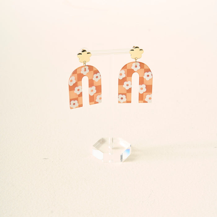 The Darling Effect's Orange Game Day Acrylic Earrings feature gold cloud-shaped studs and arched, peach dangles with white daisy patterns, displayed on a clear acrylic stand against a light background.