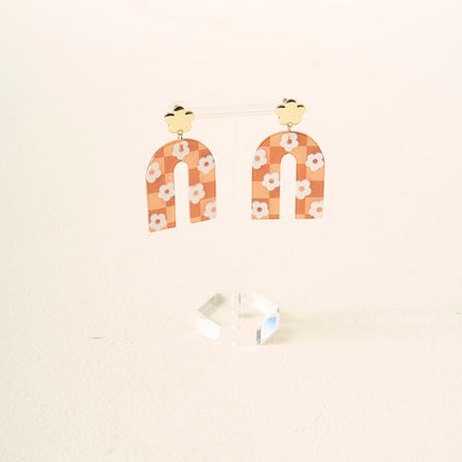 The Darling Effect's Orange Game Day Acrylic Earrings feature gold cloud-shaped studs and arched, peach dangles with white daisy patterns, displayed on a clear acrylic stand against a light background.