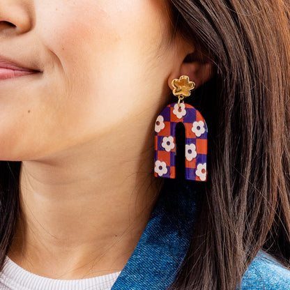 Close-up of a woman wearing The Darling Effect’s Orange Purple Game Day Acrylic Earrings—arch-shaped with a floral pattern in orange and purple, attached to a gold-colored flower stud. Perfect for showing off your school colors on game day!.