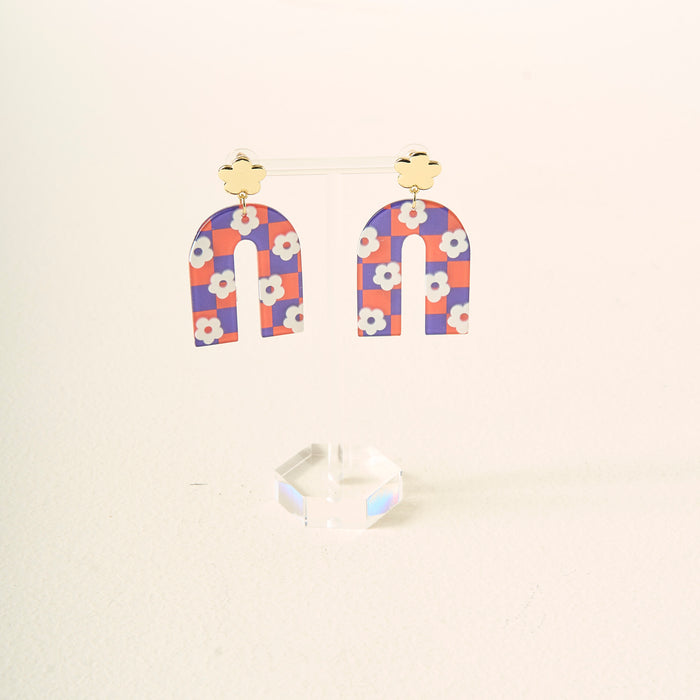 The Darling Effect's Orange Purple Game Day Acrylic Earrings feature gold clover studs and arch dangles in an orange and purple checkered pattern with white flowers, perfect for showing school spirit, displayed on a clear acrylic stand.