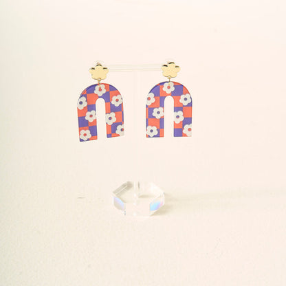 The Darling Effect's Orange Purple Game Day Acrylic Earrings feature gold clover studs and arch dangles in an orange and purple checkered pattern with white flowers, perfect for showing school spirit, displayed on a clear acrylic stand.