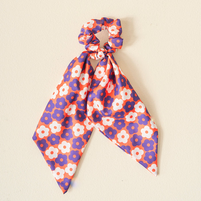 The Darling Effect's Orange Purple Game Day Hair Scarf with Scrunchie, featuring floral prints on silky fabric, makes a perfect team spirit accessory. Displayed against a light beige background.