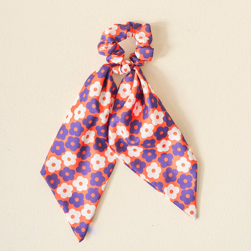 The Darling Effect's Orange Purple Game Day Hair Scarf with Scrunchie, featuring floral prints on silky fabric, makes a perfect team spirit accessory. Displayed against a light beige background.