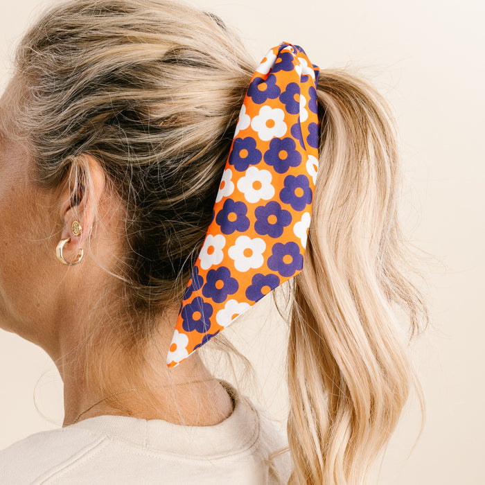 A woman with blonde hair in a ponytail wears The Darling Effect's Orange Purple Game Day Hair Scarf with Scrunchie, featuring a floral pattern. She pairs it with small hoop earrings and a light top against a plain beige background.