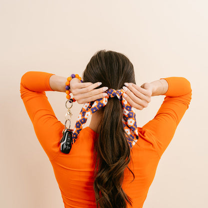 A woman in an orange top faces away, tying her long brown hair with The Darling Effect's Orange Purple Game Day Hair Scarf with Scrunchie. On her wrist is a beaded keychain bracelet holding car keys, set against a plain beige background.