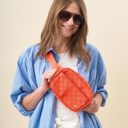 A smiling woman wears sunglasses, a blue striped shirt over a white top, and holds The Darling Effect’s Orange Urban Check Belt Bag against a plain background.
