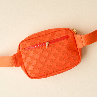 The Darling Effect’s Orange Urban Check Belt Bag features a front zipper pocket, matching adjustable strap, and vegan leather—making it a trendy accessory for any outfit. Displayed on a light beige surface.