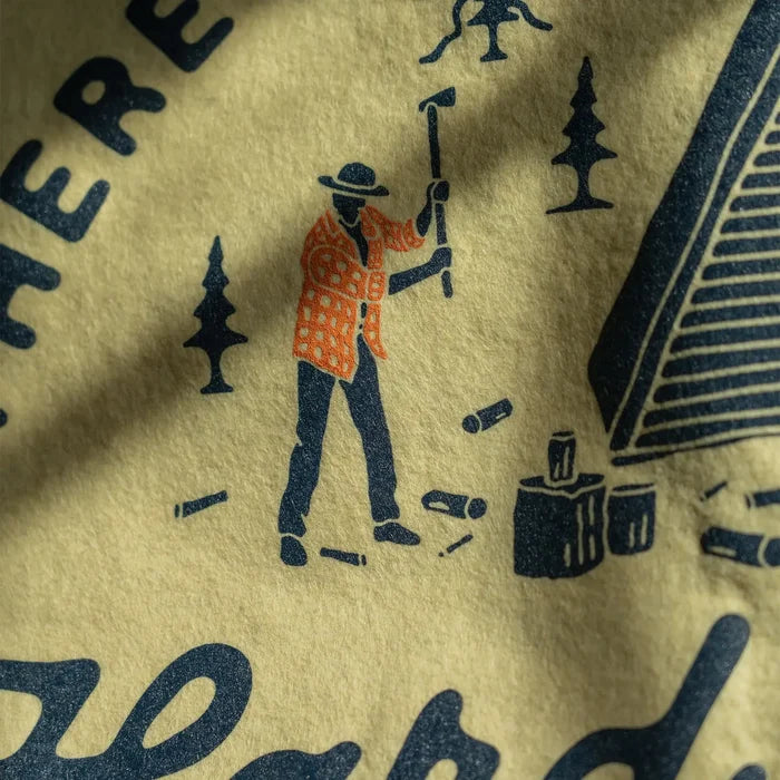 Person in plaid chopping wood by rustic cabin with trees, logs, and Oxford Pennant “No Substitute for Hard Work” flag nearby.